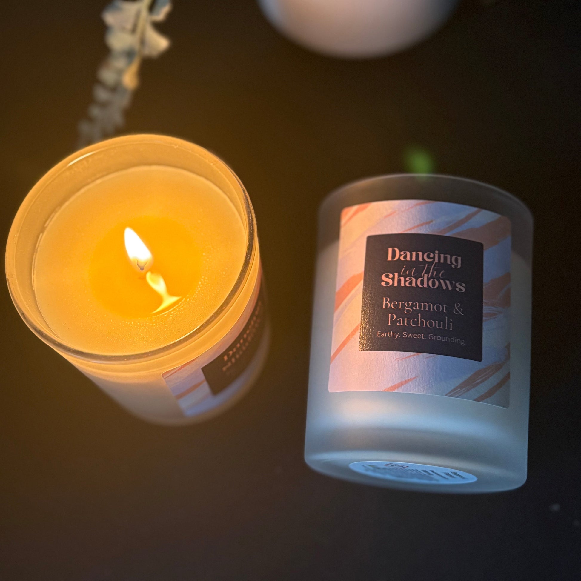 Candle with a lit flame and 'Dancing in the Shadows' candle lying next to it on a flat surface.