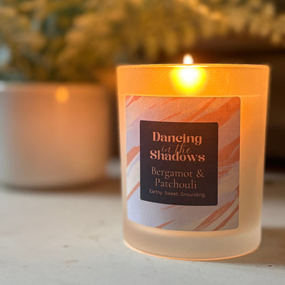 Lit candle labeled 'Dancing in the Shadows' with bergamot and patchouli scent on a surface with a blurred plant background.