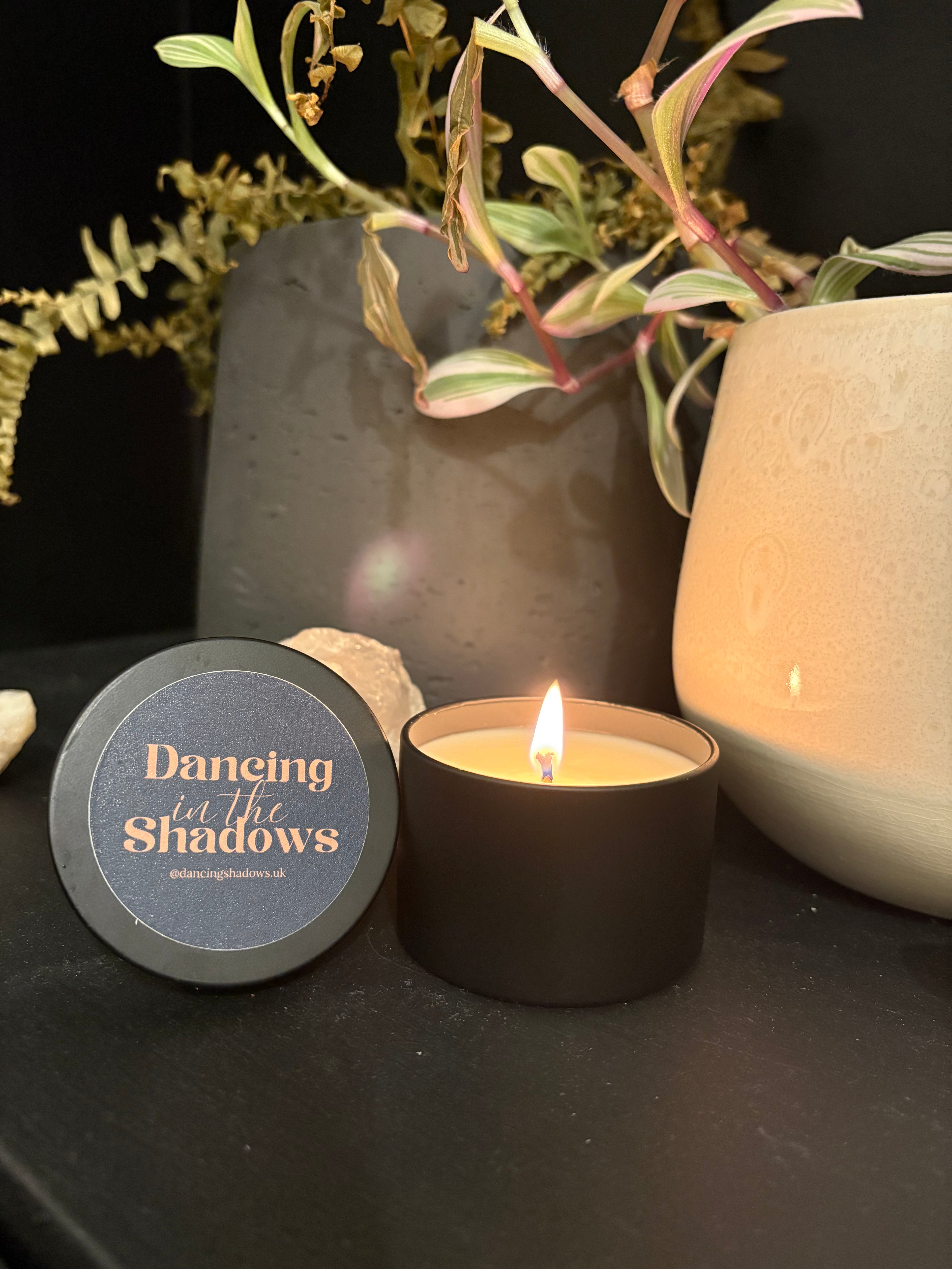 Candle with a small round container labeled 'Dancing in the Shadows' on a dark surface with greenery.